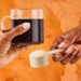 a cup of coffee in a hand and a scoop of collagen powder in another hand with an orange background