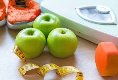 A scale, measuring tape, apples, dumbbell & running shoes sitting next to one another.