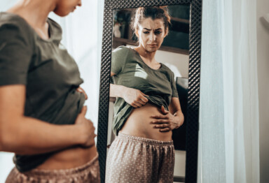 women looking in the mirror holding her stomach as if she is looking at her bloated stomach